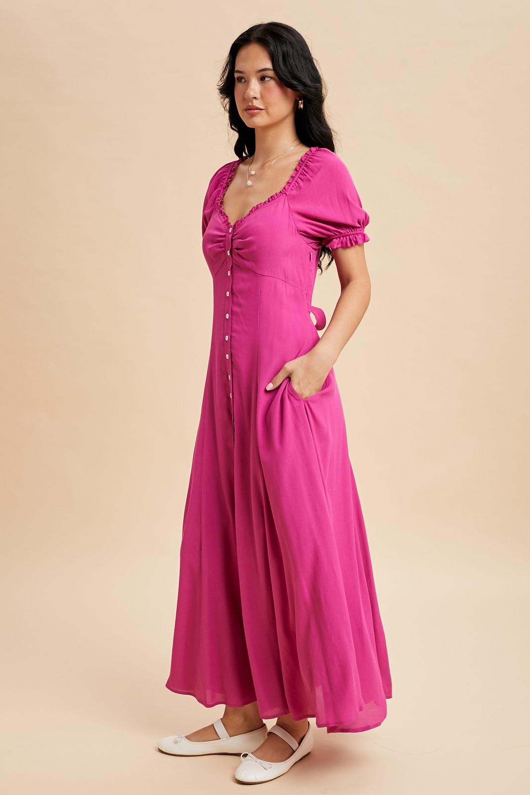 woman wearing a bright pink short sleeve maxi dress with buttons and pockets, standing in profile against a neutral background