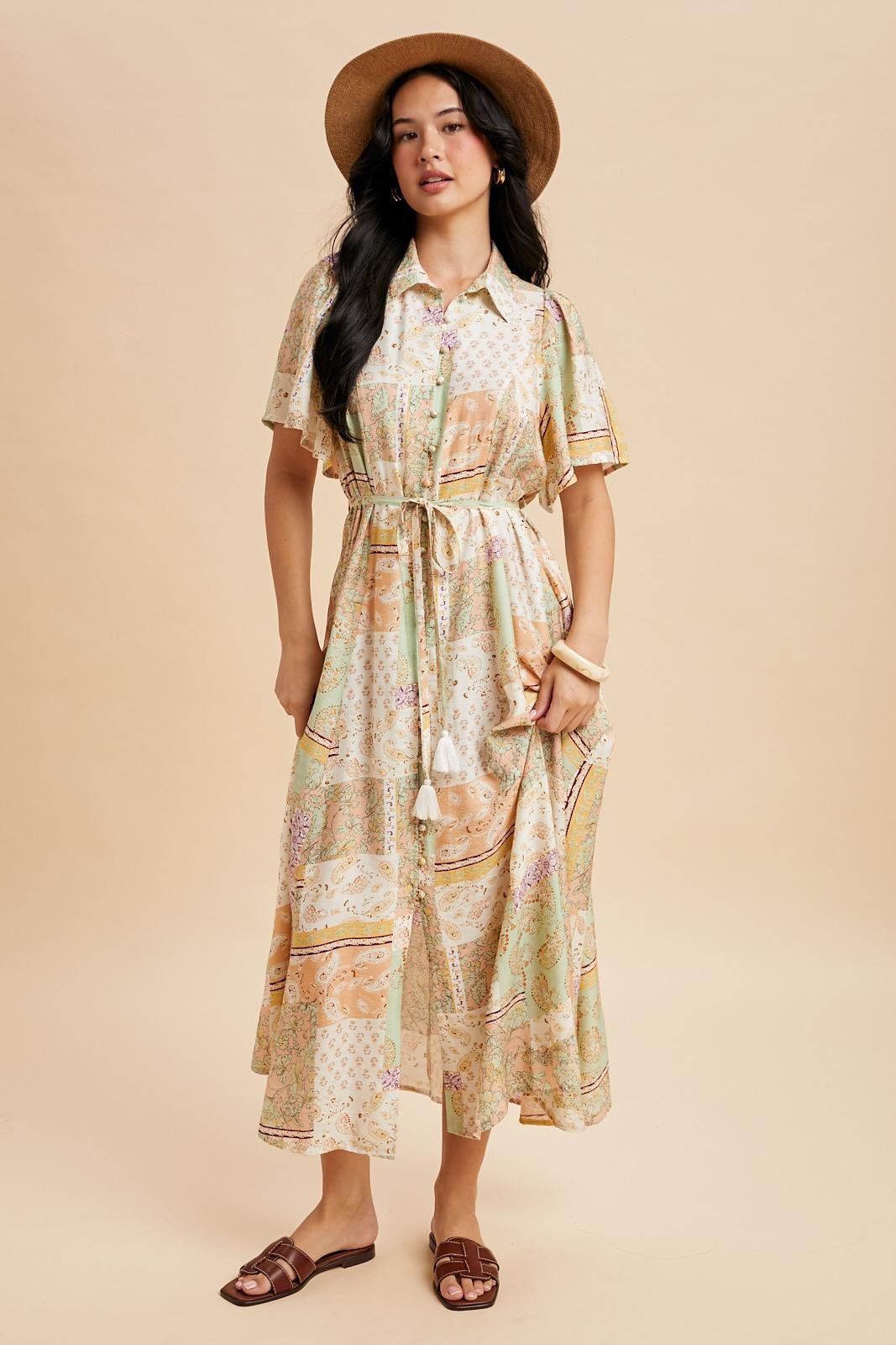 woman wearing a pastel patterned midi dress with short sleeves and a tie belt, accessorized with a hat and sandals