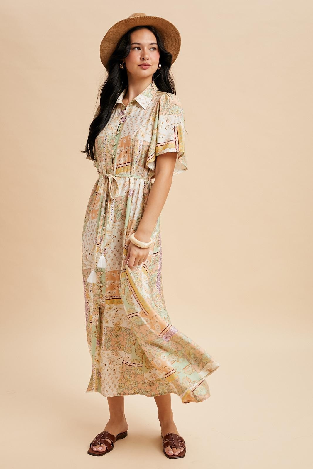 woman wearing a patterned maxi dress with short sleeves, cinched waist, and tassel details, paired with a wide-brimmed hat