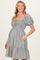 woman wearing a black and white checkered maternity dress with puff sleeves and a tiered hem, smiling and posing with her hand on her hip
