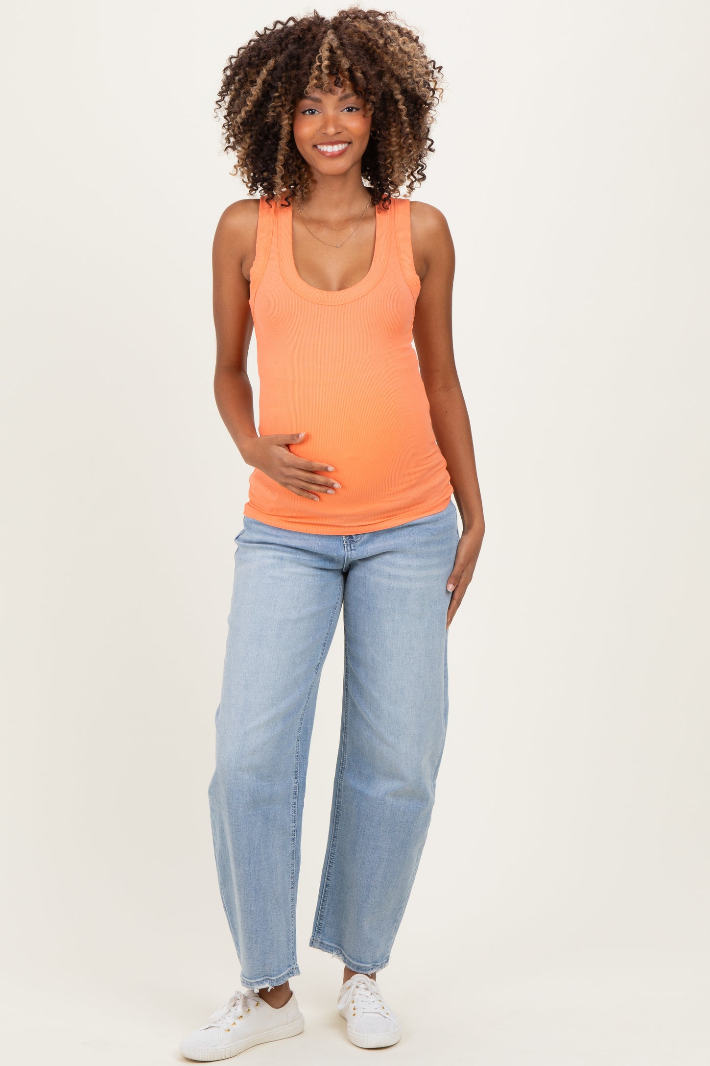 woman wearing an orange ribbed maternity tank top and light blue jeans, gently cradling her baby bump