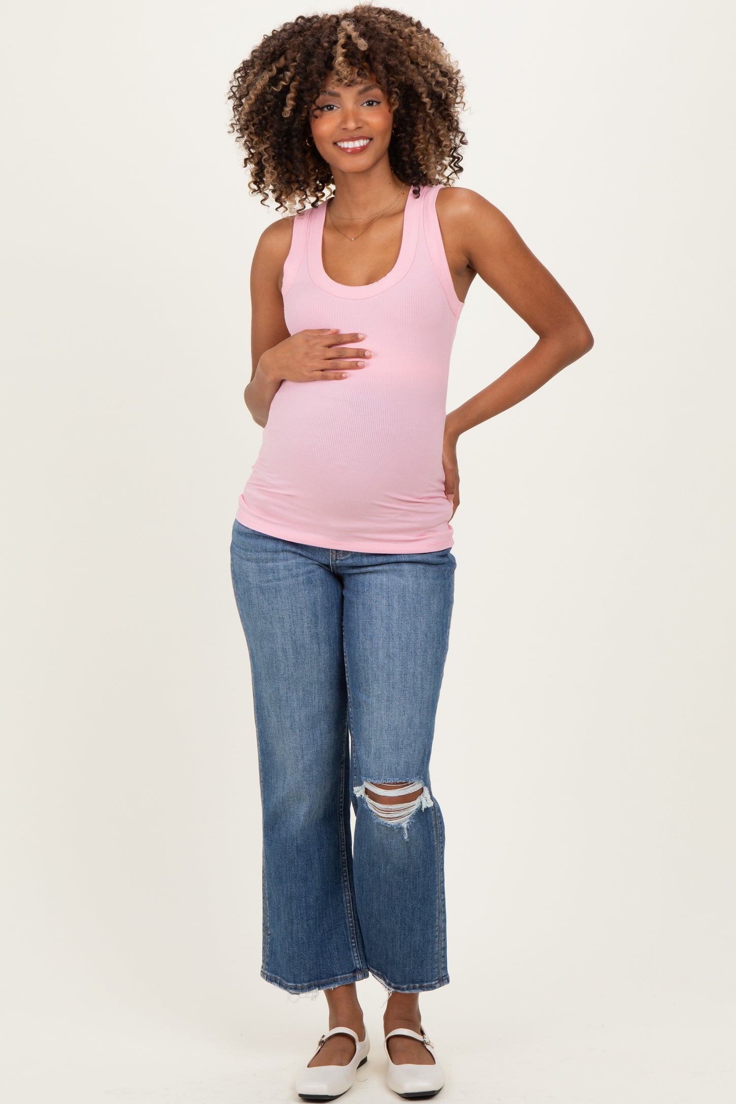 woman wearing a light pink ribbed maternity tank top and distressed flared jeans, holding her baby bump