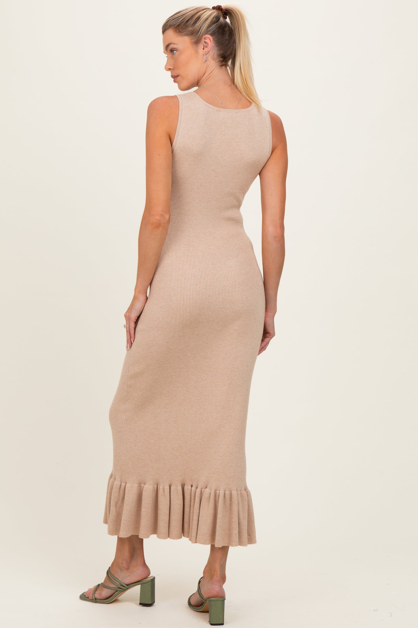 woman wearing a beige ribbed maternity dress with a ruffled hem, showing her back and styled with green heels