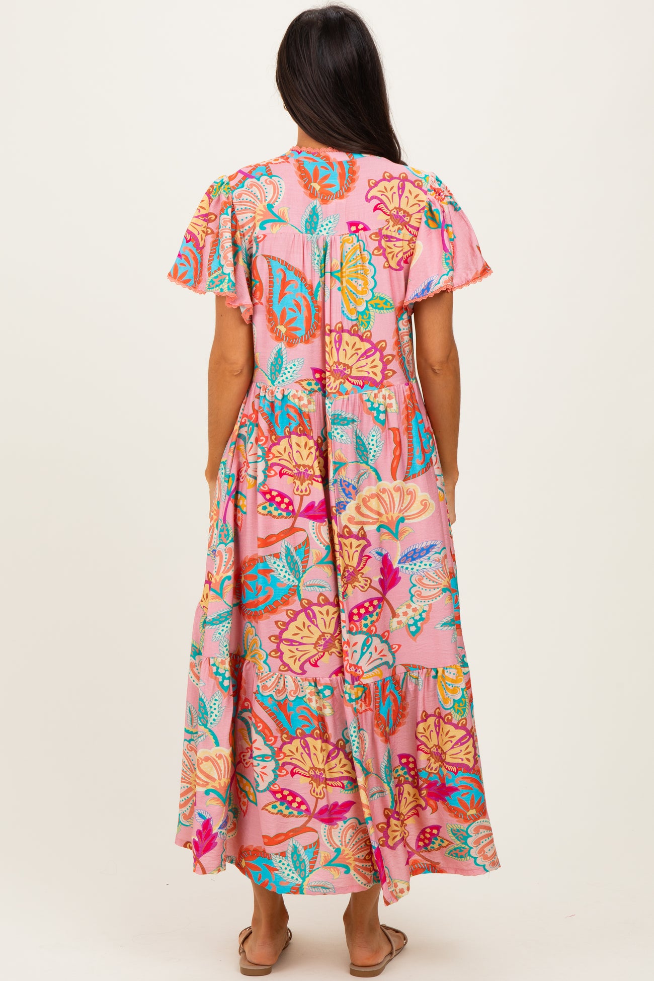 woman wearing a colorful floral maxi dress with short sleeves, featuring a pink background and tiered hemline, shown from the back