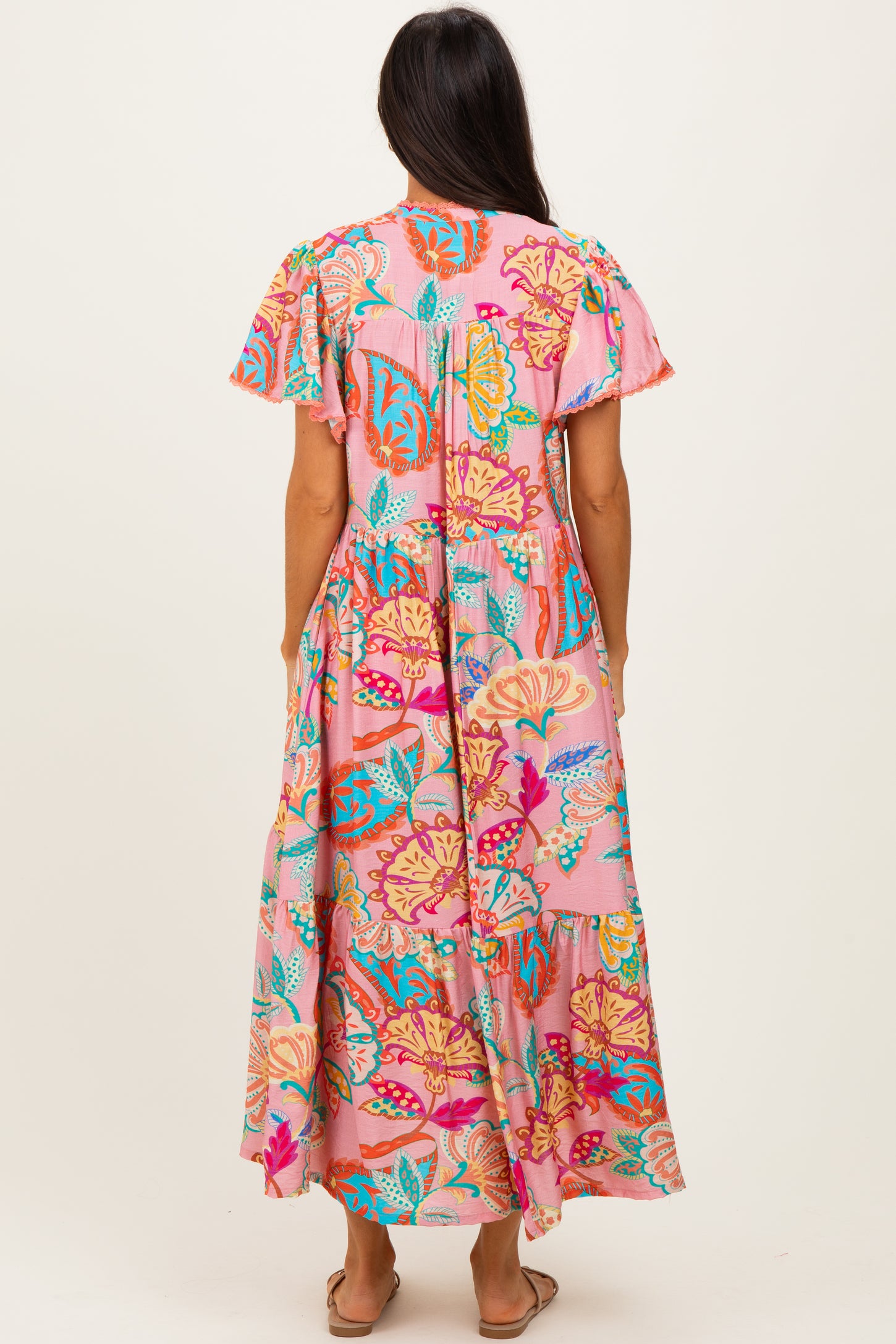 woman wearing a colorful floral maxi dress with short sleeves, featuring a pink background and tiered hemline, shown from the back