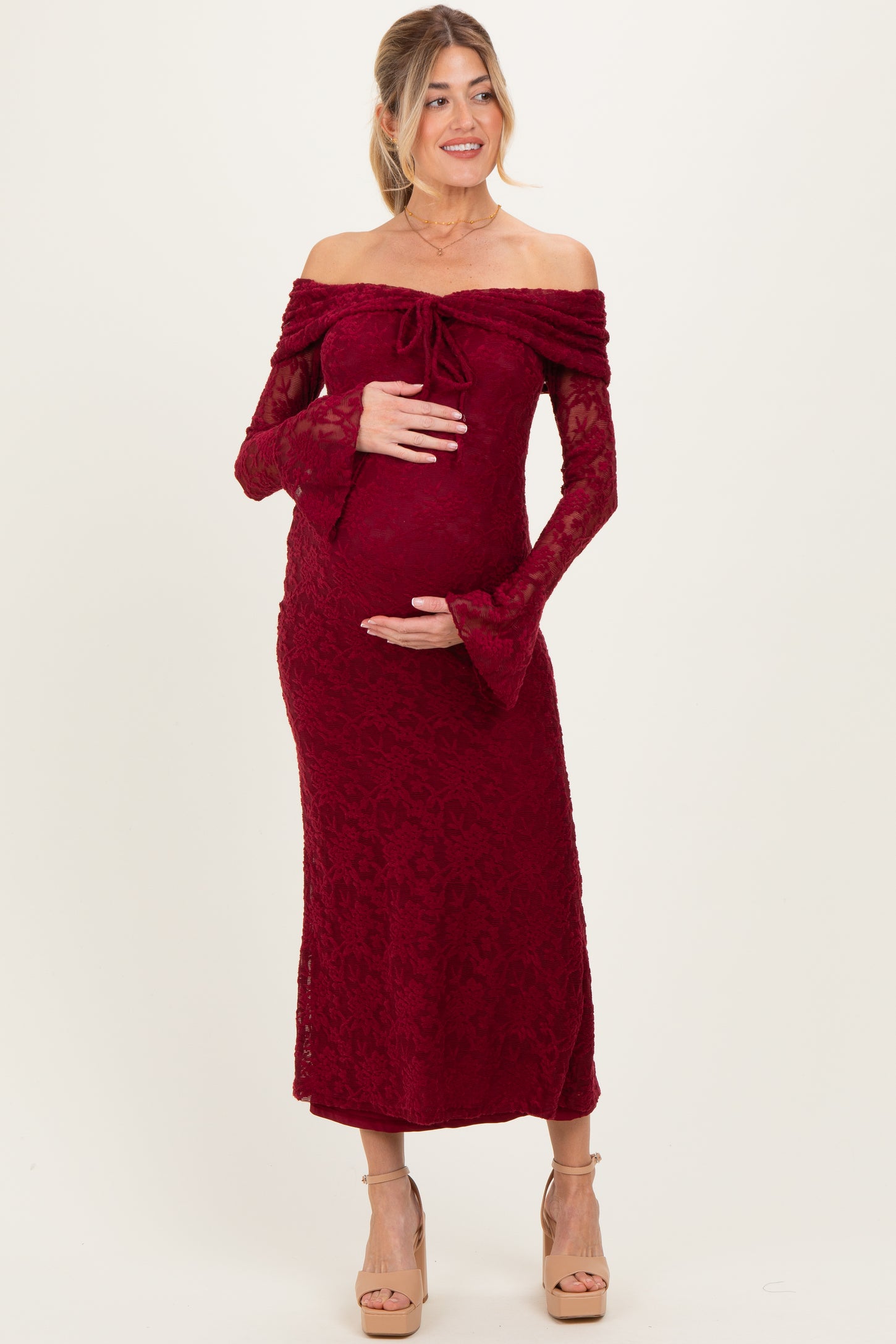woman wearing a burgundy off-the-shoulder lace maternity dress with fitted waist and bell sleeves, holding her baby bump