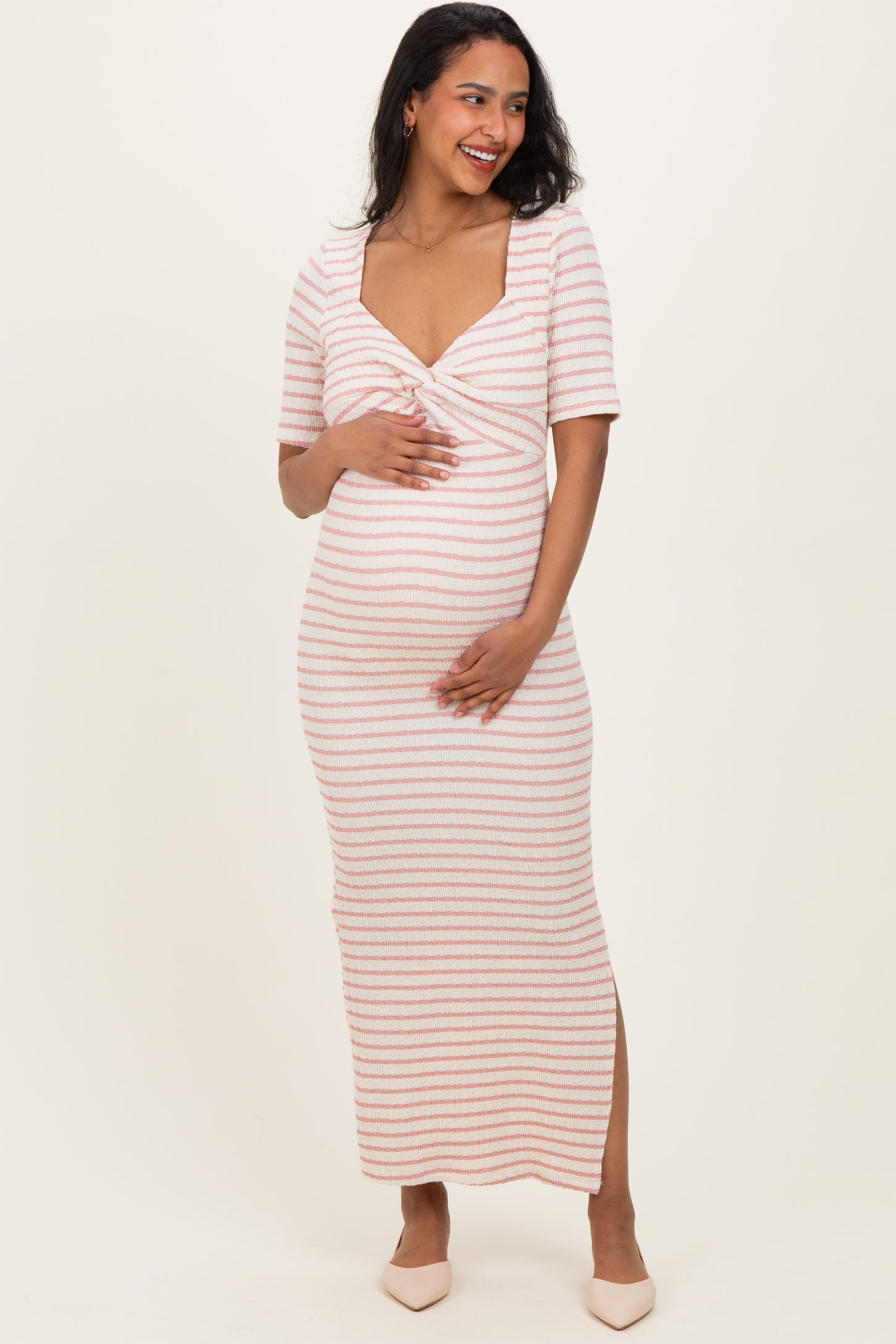 woman wearing a pink and white striped maternity maxi dress with a knotted front and side slit, holding her baby bump