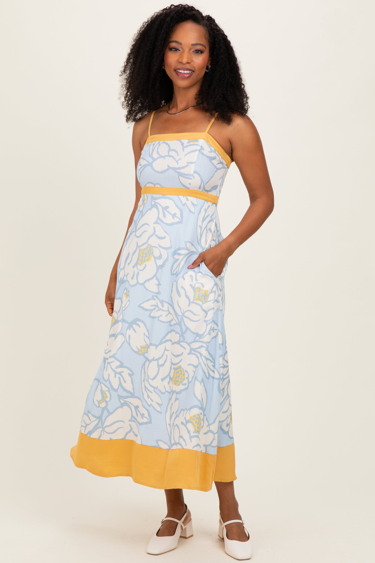 woman wearing a light blue floral maxi dress with yellow accents and pockets, standing against a plain background