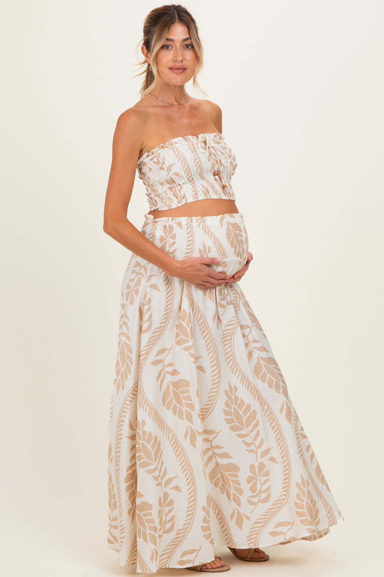 woman wearing a cream and beige floral maternity two-piece outfit with a strapless top and long skirt, holding her baby bump