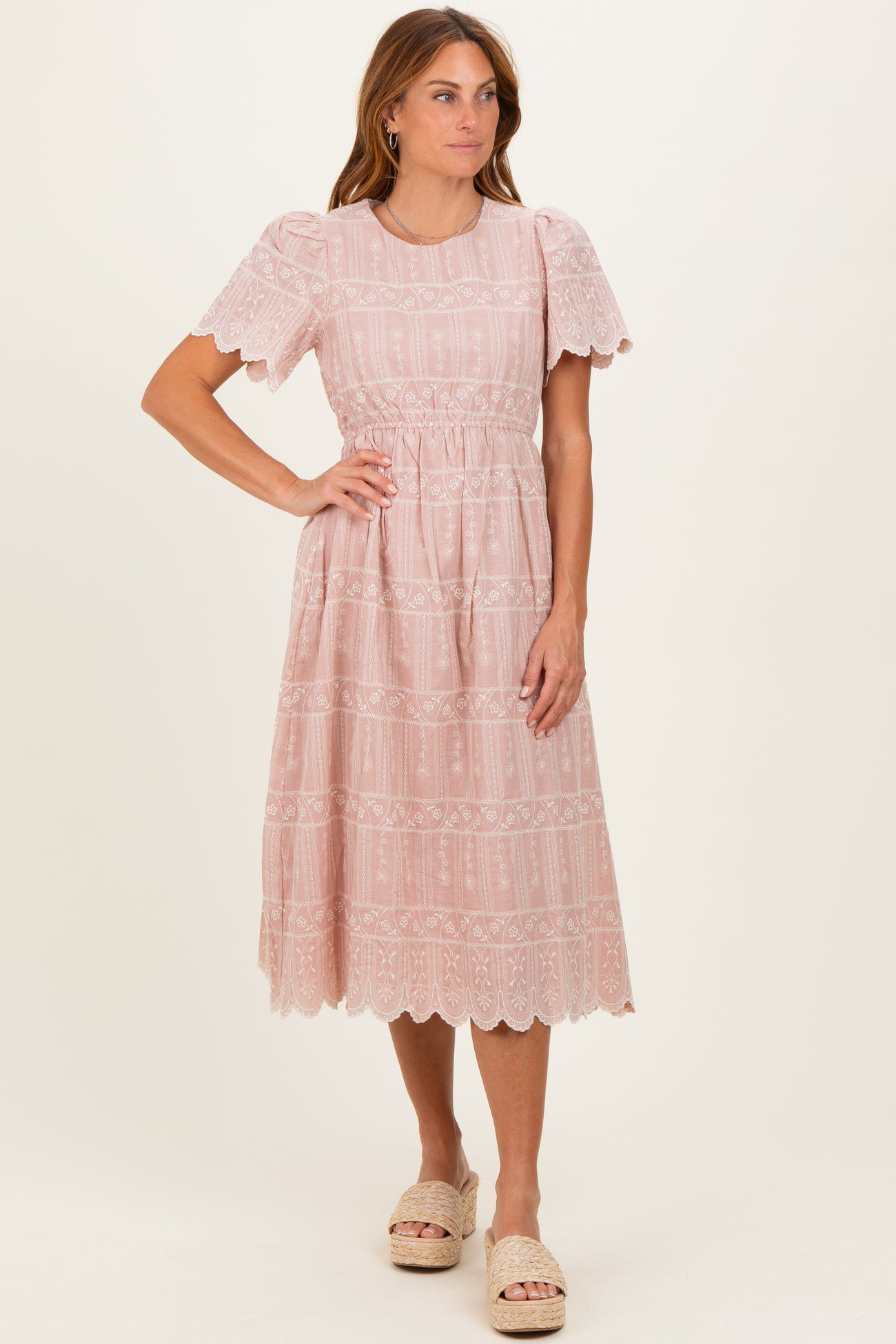 woman wearing a light pink embroidered midi dress with short ruffled sleeves and a fitted waist, standing on a neutral background