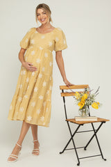 woman wearing a yellow floral maternity dress with puff sleeves and a visible baby bump, standing next to a chair