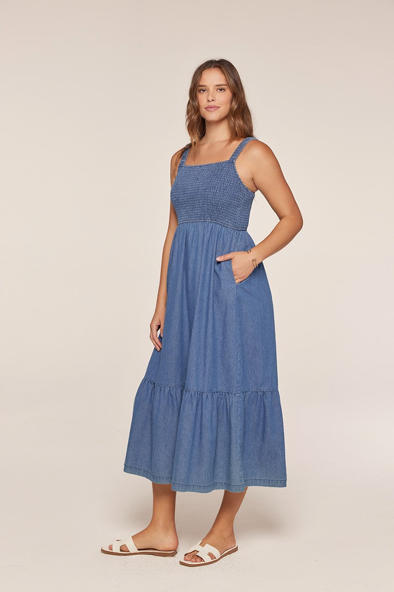 woman wearing a denim maxi dress with smocked bodice and tiered skirt, standing with one hand in pocket