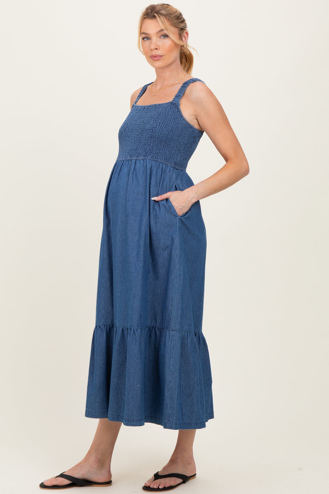 woman wearing a blue denim maternity maxi dress with smocked bodice and tiered skirt, hands in pockets, showing baby bump