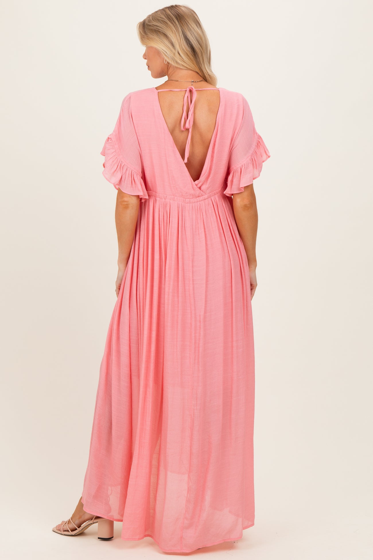 woman wearing a pink backless maxi maternity dress with ruffle sleeves and a tie at the neck