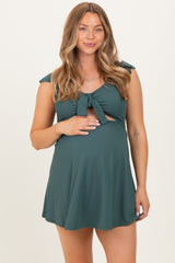 woman wearing a dark green maternity dress with tie straps and cut-out details, holding her baby bump