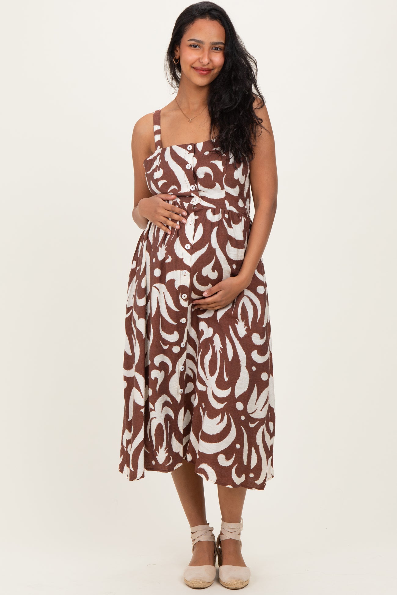 woman wearing a lightweight brown and white patterned maternity dress, gently cradling her baby bump