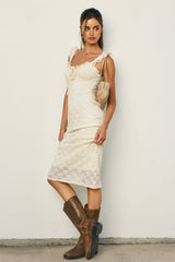 woman wearing a white lace dress with spaghetti straps and brown knee-high boots, posing against a neutral background