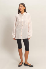 woman wearing a light pink oversized button-up shirt with two chest pockets and black leggings, standing with one foot forward