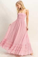woman wearing a pink maxi dress with a smocked bodice and ruffled hem, standing and holding the skirt with both hands