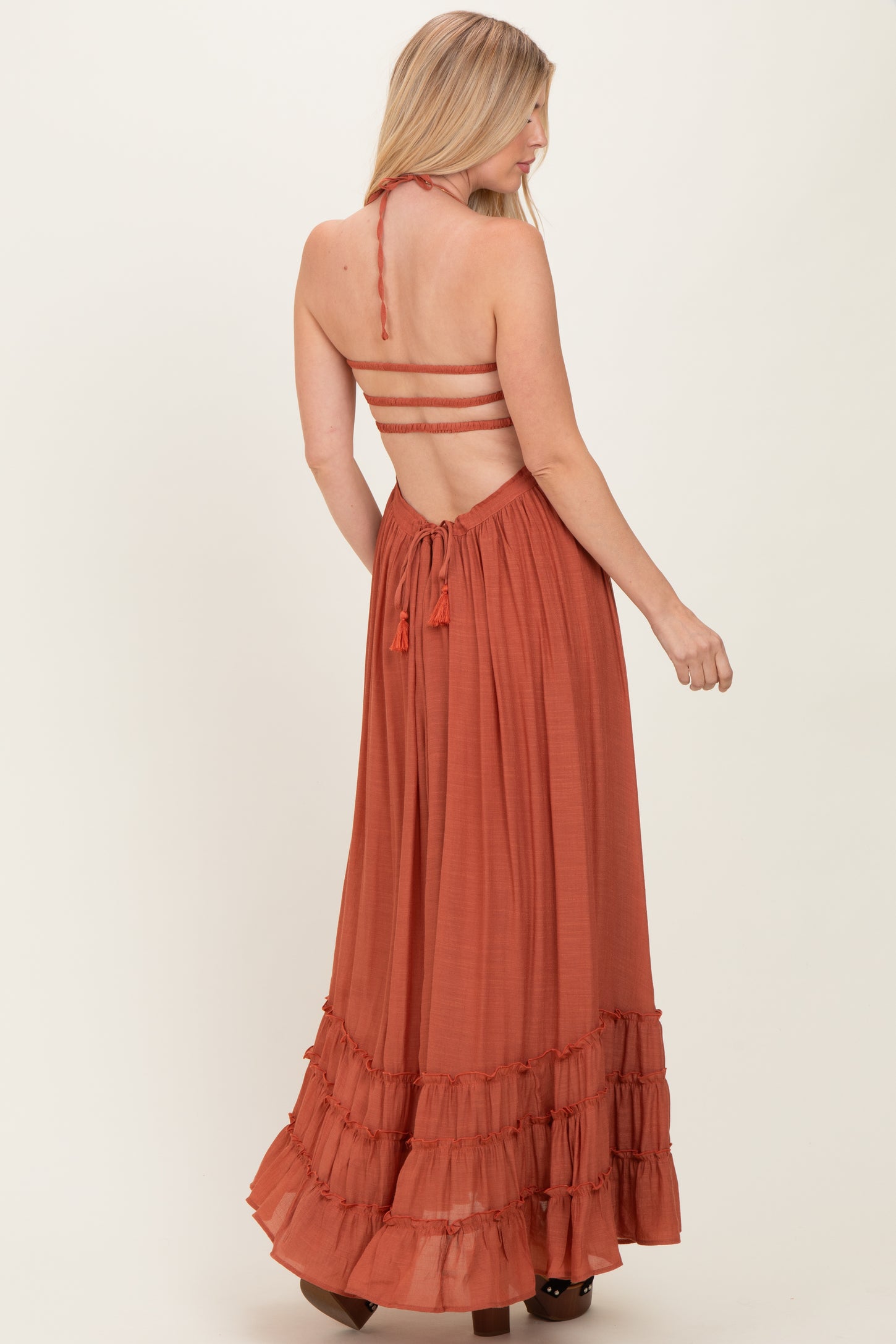 woman wearing a rust-colored maxi dress with a back tie and ruffle details, styled with spaghetti straps