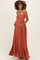 woman wearing an rust-colored maternity maxi dress with a smocked bodice and ruffle hem, holding her baby bump