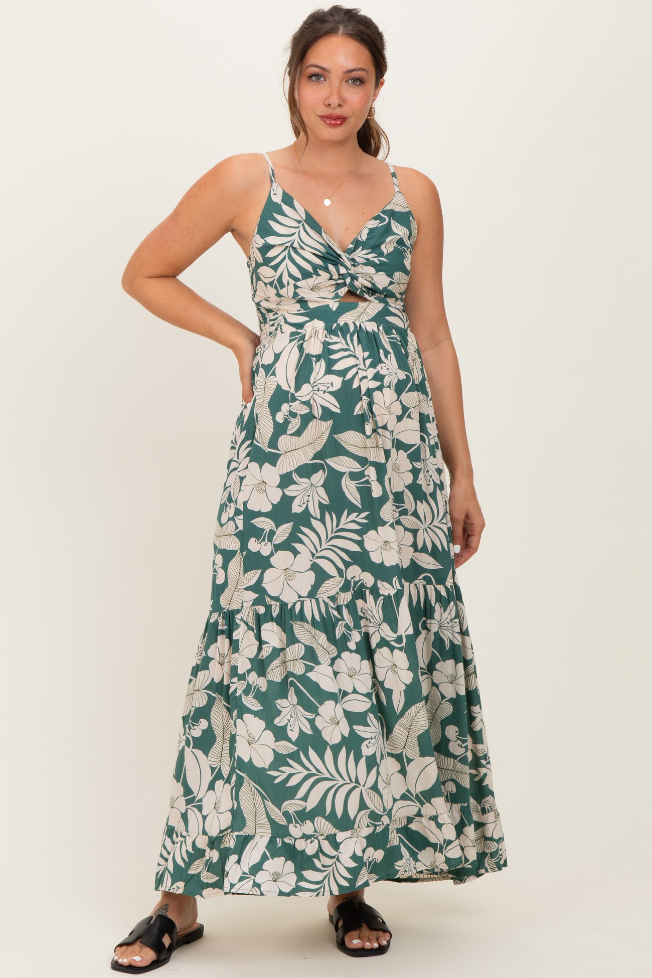 woman wearing a green floral print maternity maxi dress with a cinched waist and spaghetti straps, showing off her baby bump