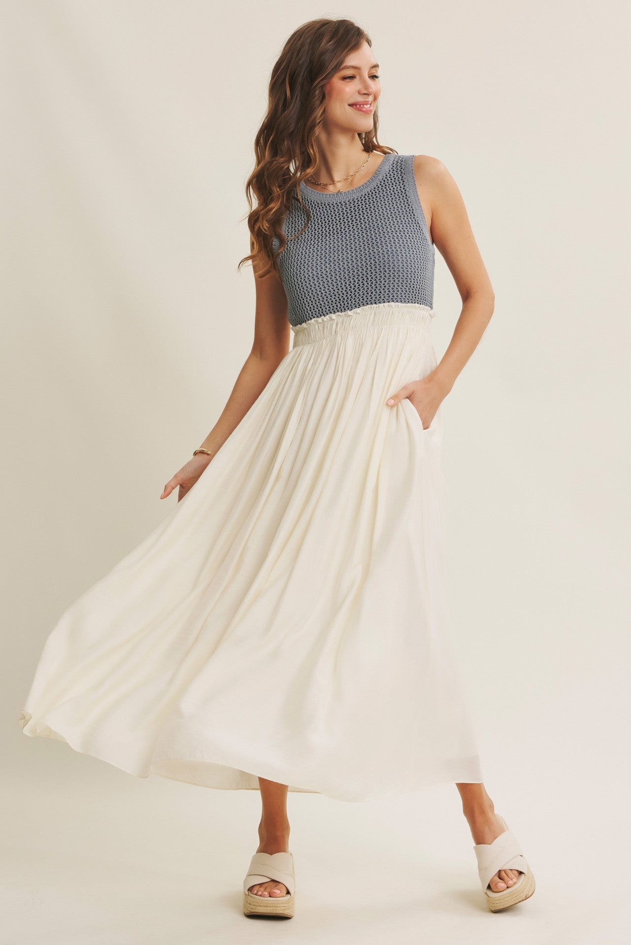 woman wearing a sleeveless blue knit and white maxi dress with a flowy skirt and pockets, standing against a neutral background
