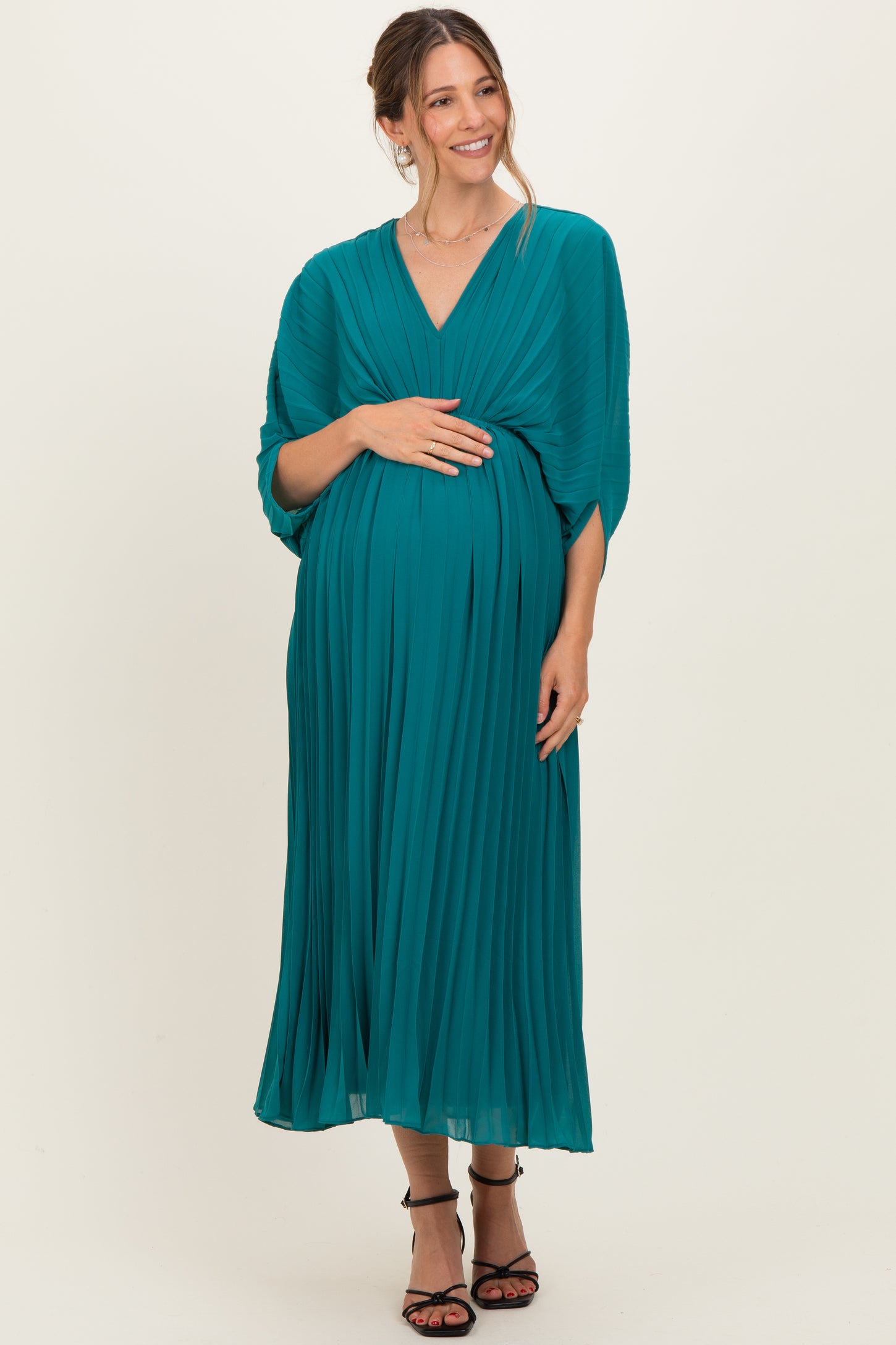 woman wearing a teal pleated maternity dress with a v-neck and 3/4 sleeves, holding her baby bump