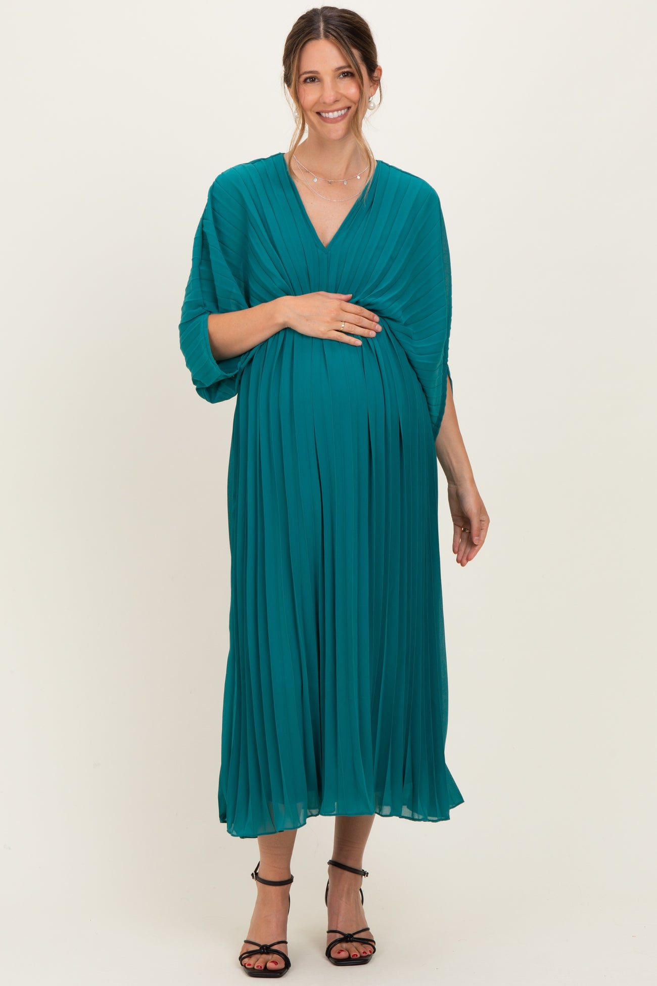 woman wearing a teal pleated maternity dress, gently cradling her baby bump