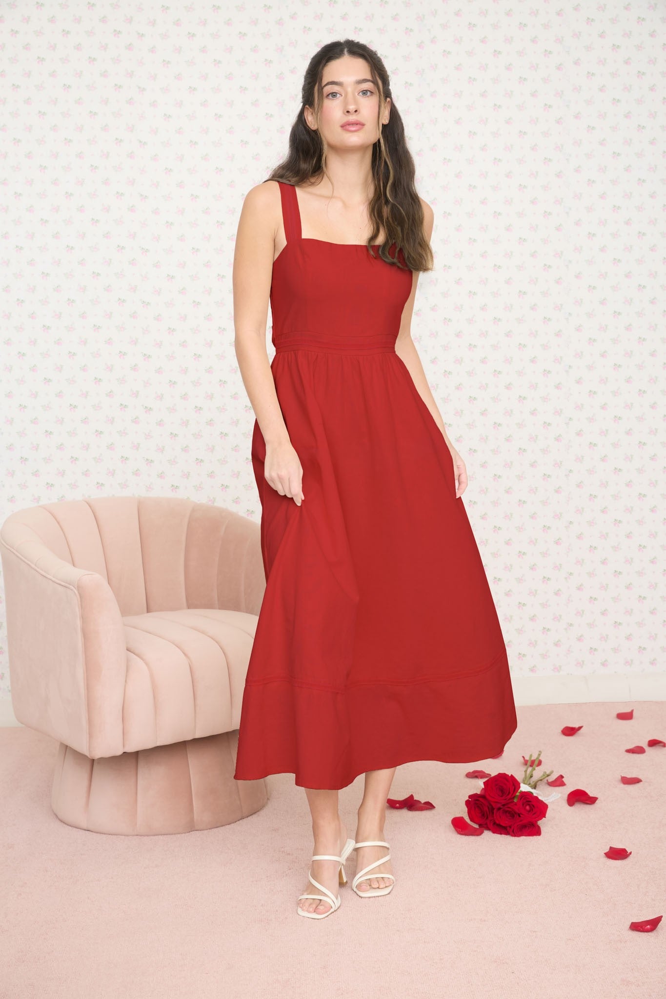 woman wearing a red midi dress with thick straps, standing in a softly decorated room with rose petals on the floor
