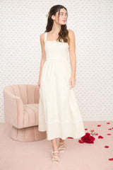 woman wearing a cream spaghetti strap maxi dress with a fitted bodice and a flowy skirt, posing next to a pink armchair
