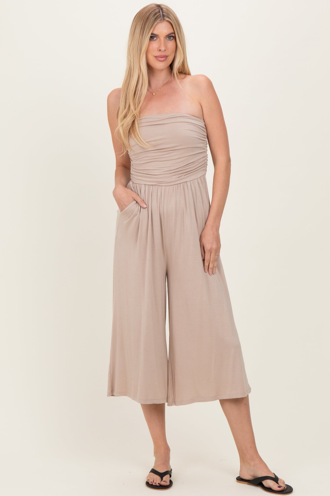 woman wearing a beige strapless jumpsuit with a ruched bodice and side pockets, standing and smiling in flip flops