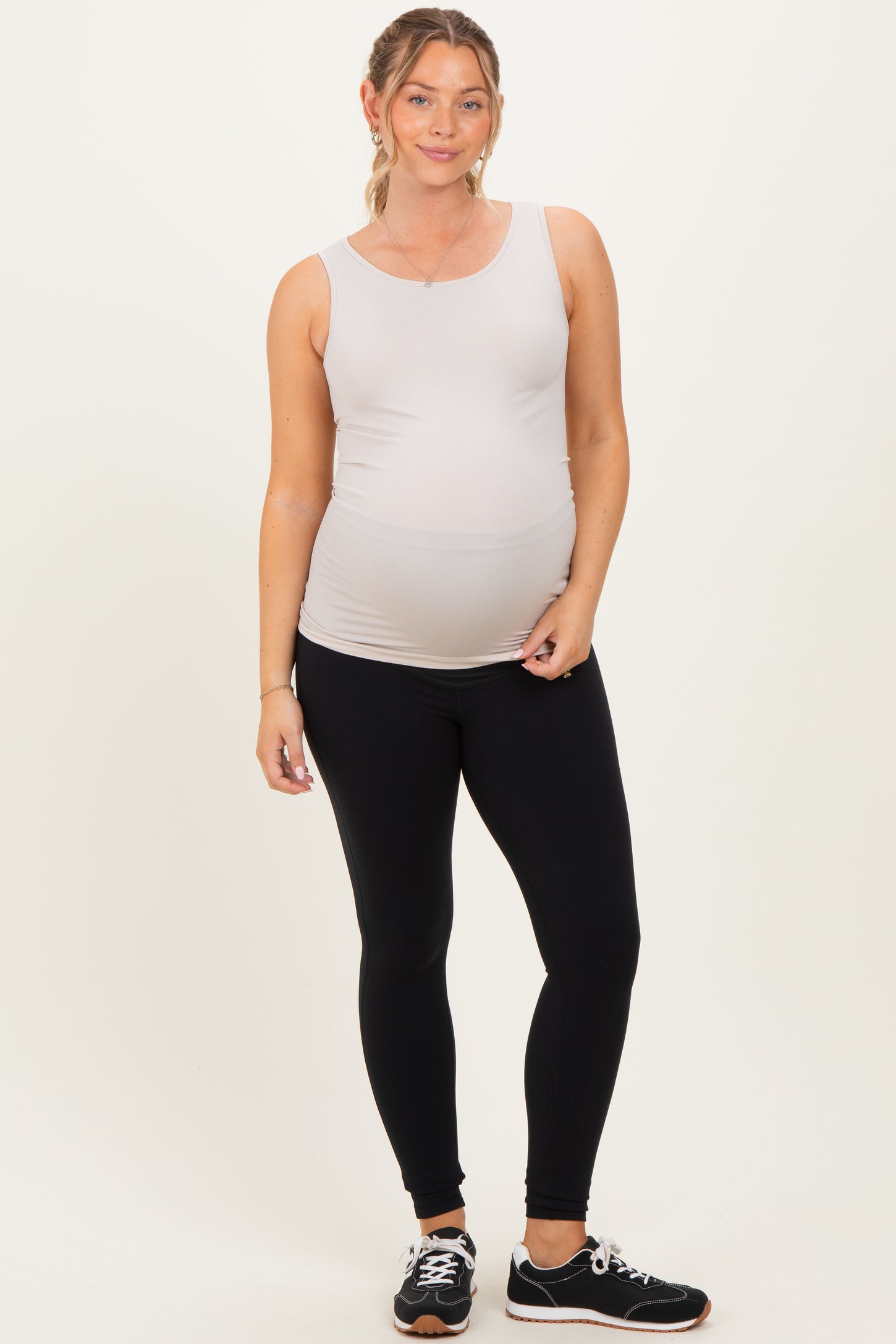 woman wearing a light gray maternity tank top and black maternity leggings, showing her baby bump