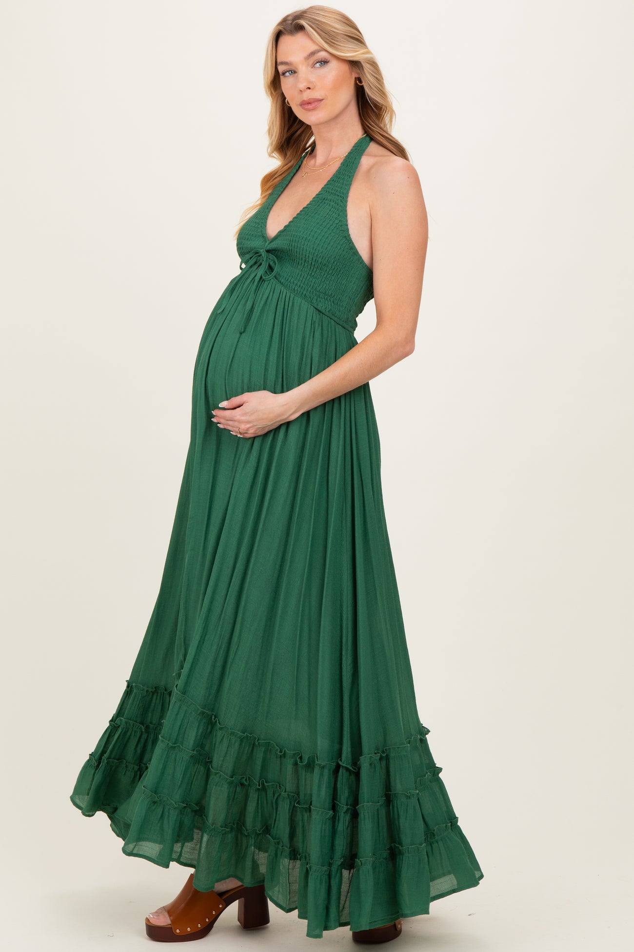 woman wearing a green halter maternity maxi dress with a ruffled hemline and holding her baby bump