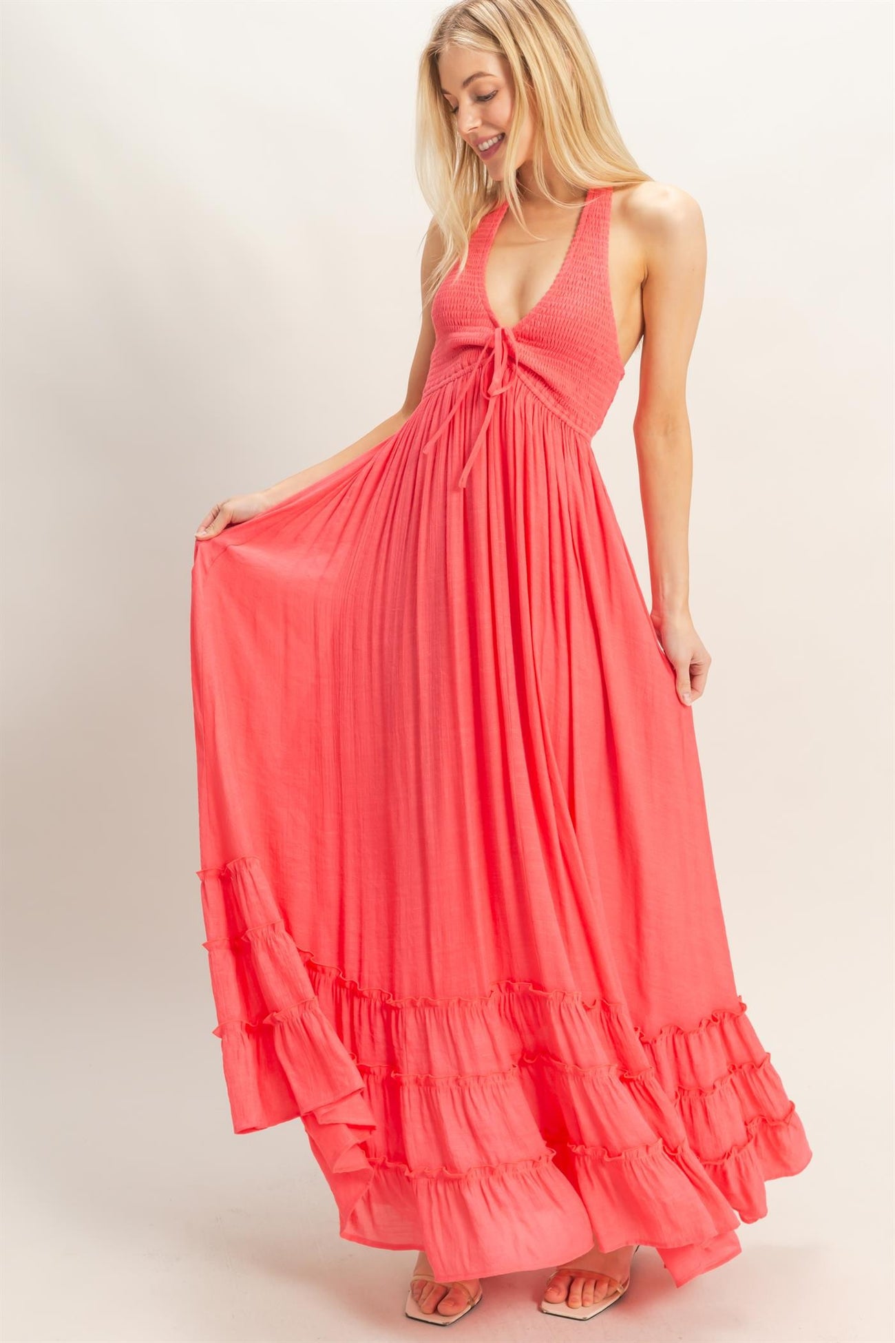 woman wearing a coral maxi dress with a deep V-neck and ruffle trim, posing and holding the hem of the dress