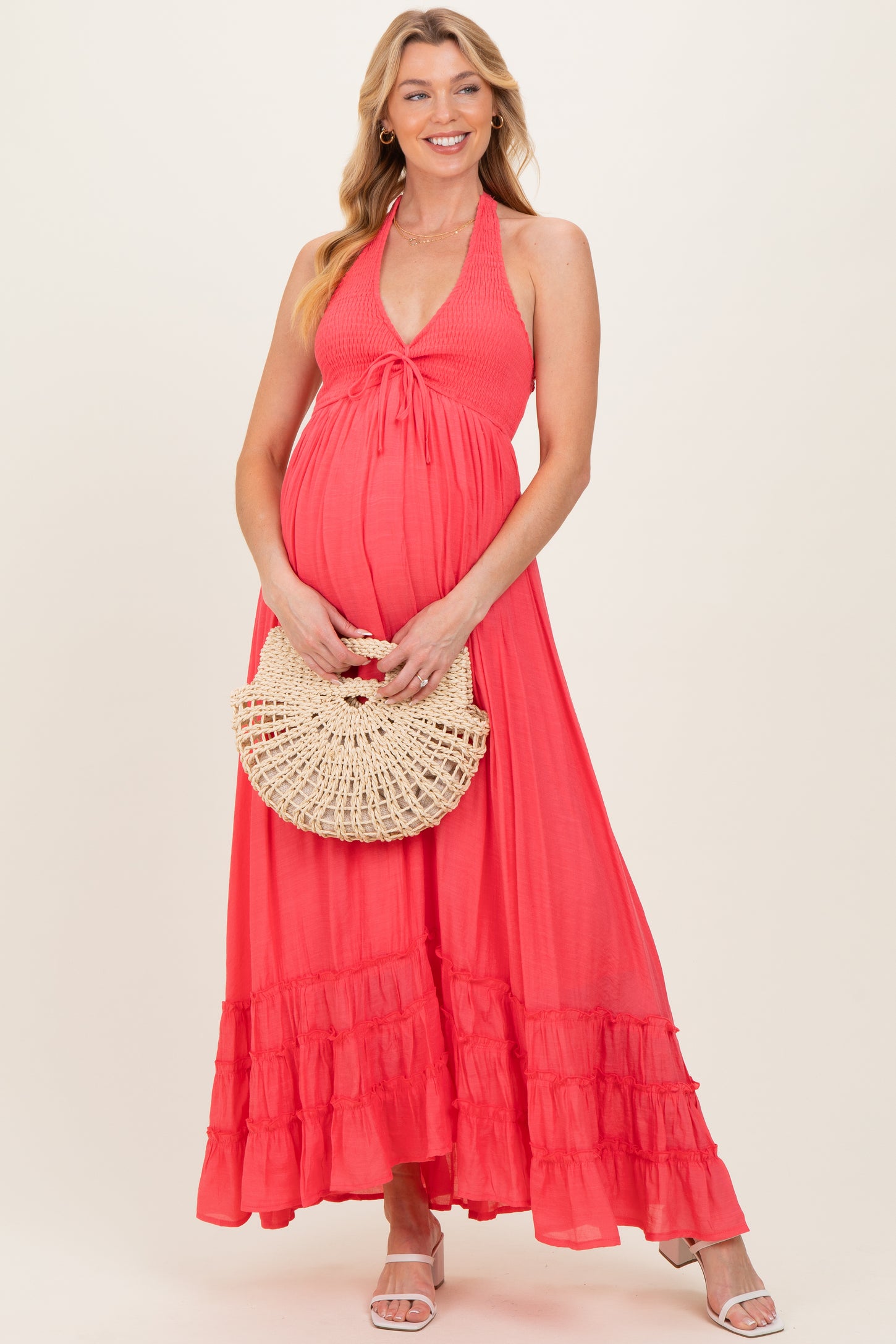 woman wearing a coral maternity maxi dress with a halter neck and tiered ruffle hem, holding a straw bag and showing her baby bump