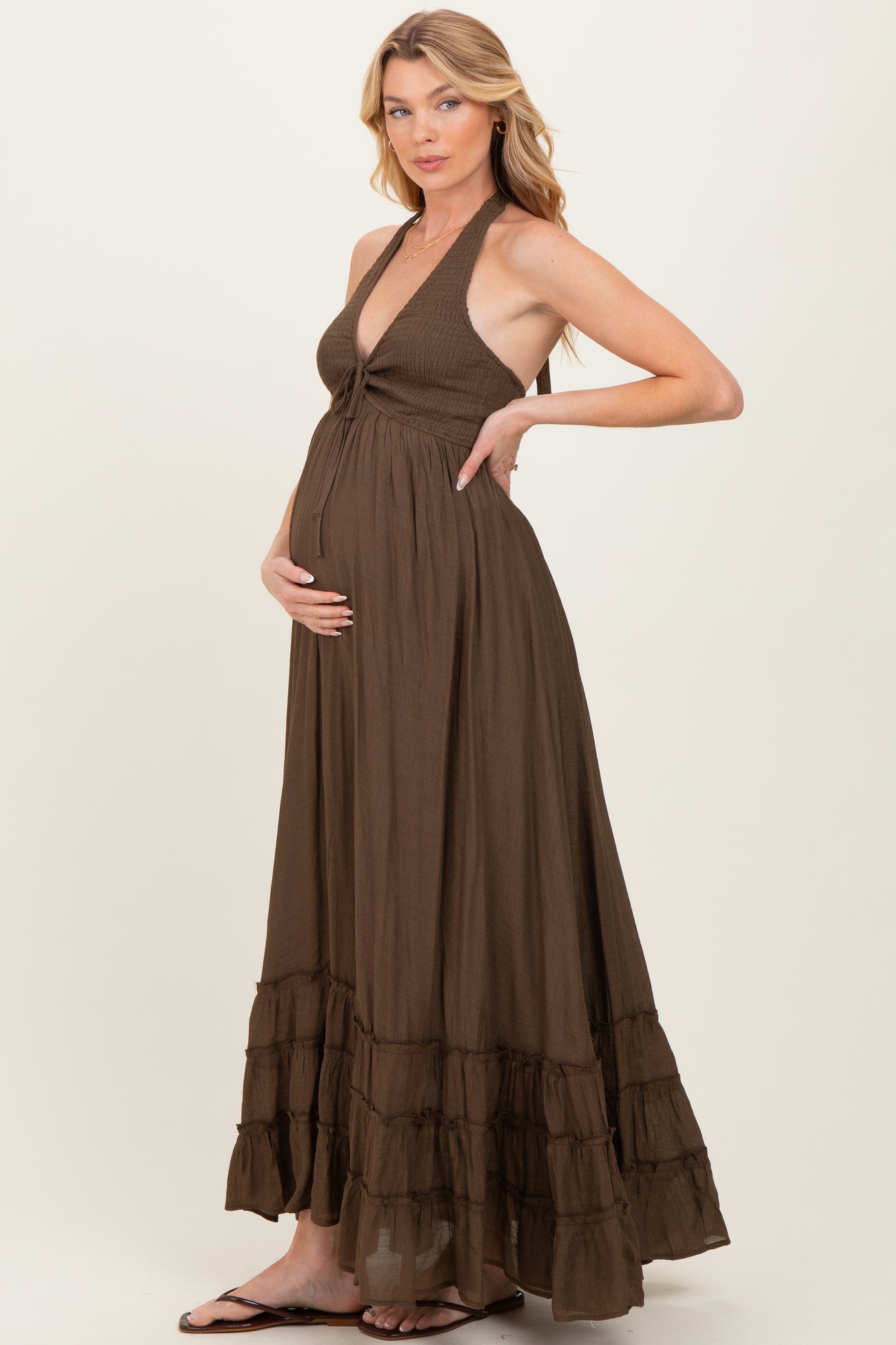 woman wearing a brown maternity maxi dress with a halter neckline and a tiered ruffle hem, holding her baby bump