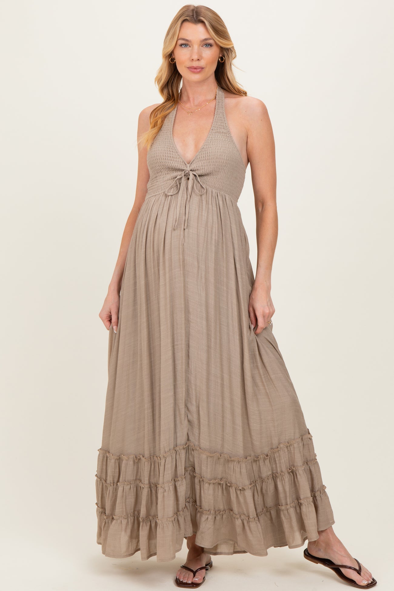 woman wearing a beige halter neck maternity maxi dress with a tie front and tiered ruffle hem, showing her baby bump