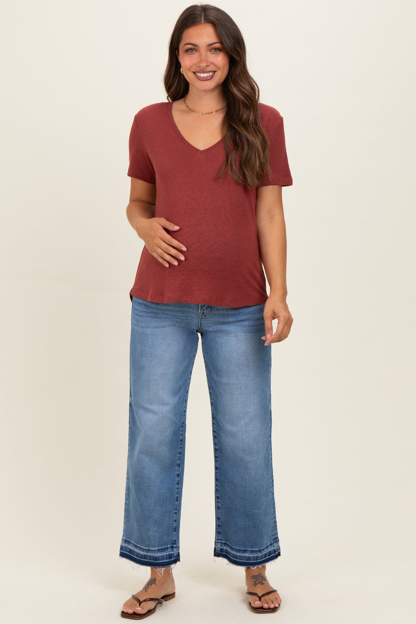 woman wearing a maroon v-neck maternity top and blue wide-leg jeans, gently holding her baby bump