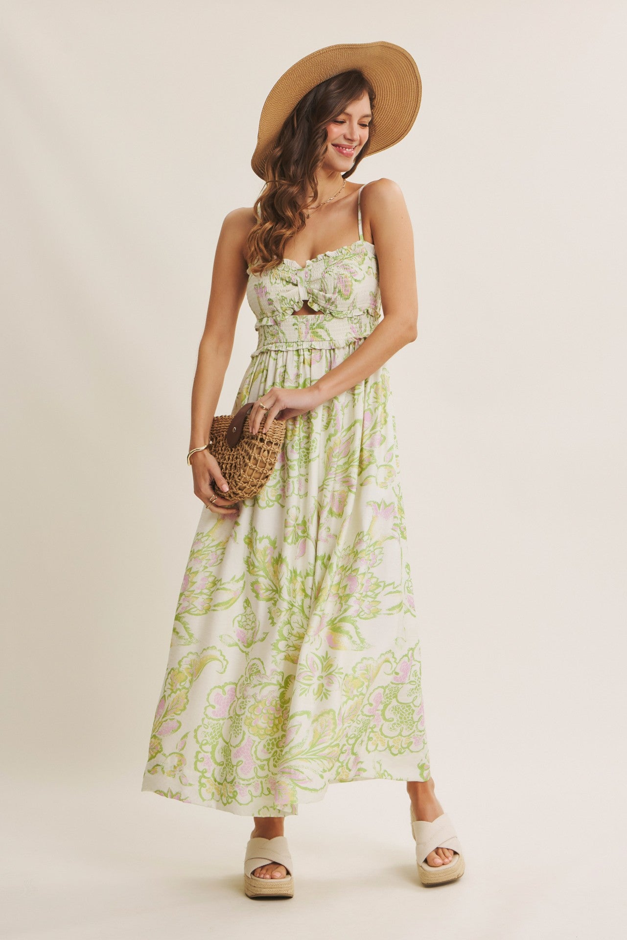 woman wearing a floral print maxi dress with spaghetti straps and a straw hat, holding a woven bag