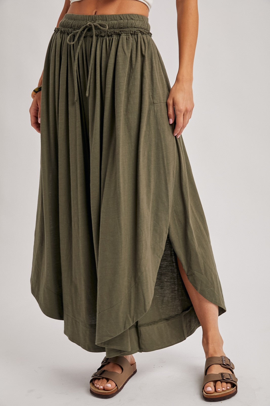 woman wearing an olive green maxi skirt with a high slit and elastic waist, standing with one hand on her hip