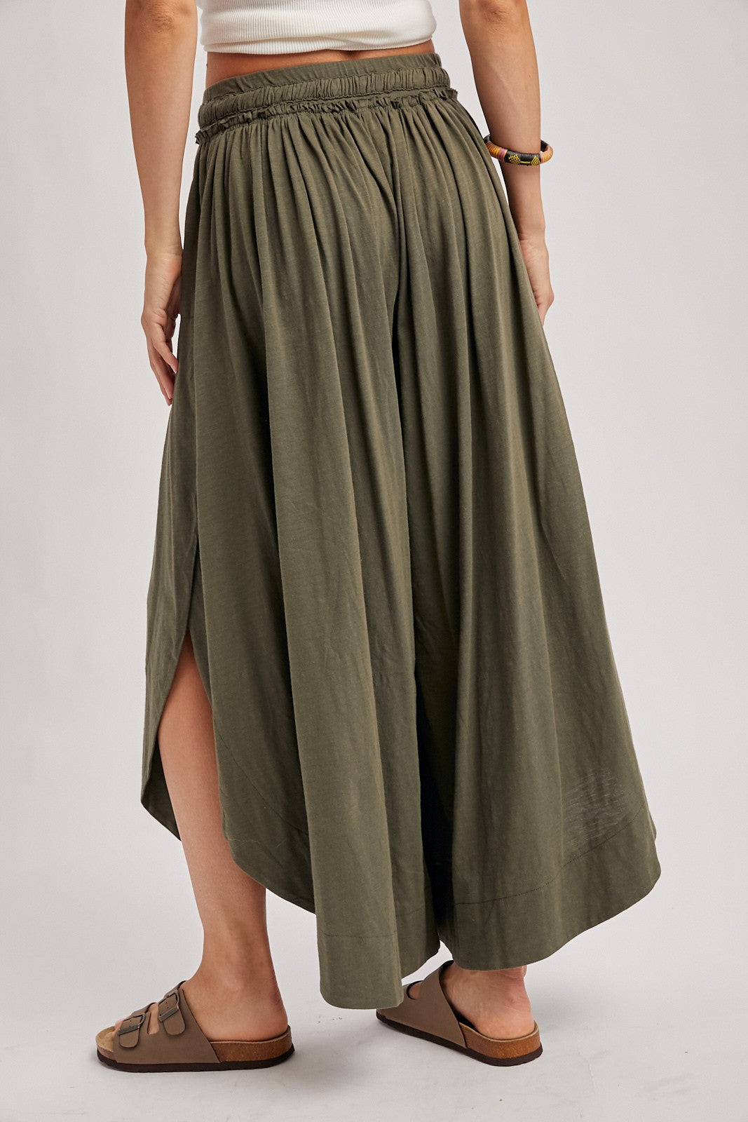 woman wearing a long olive green maxi skirt with a pleated design and side slits, showing the back view