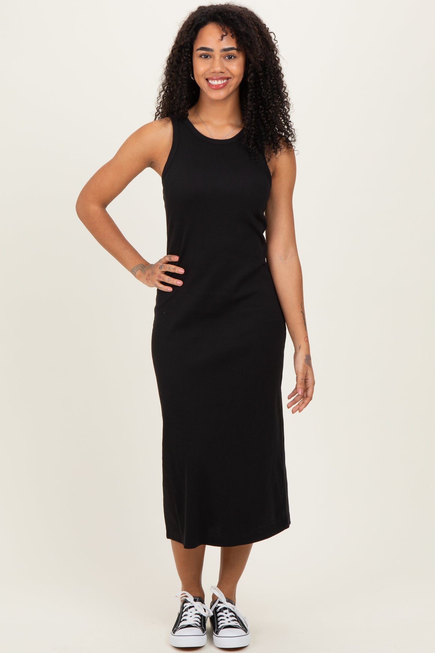 woman wearing a black sleeveless midi dress and black and white sneakers, standing with a smile