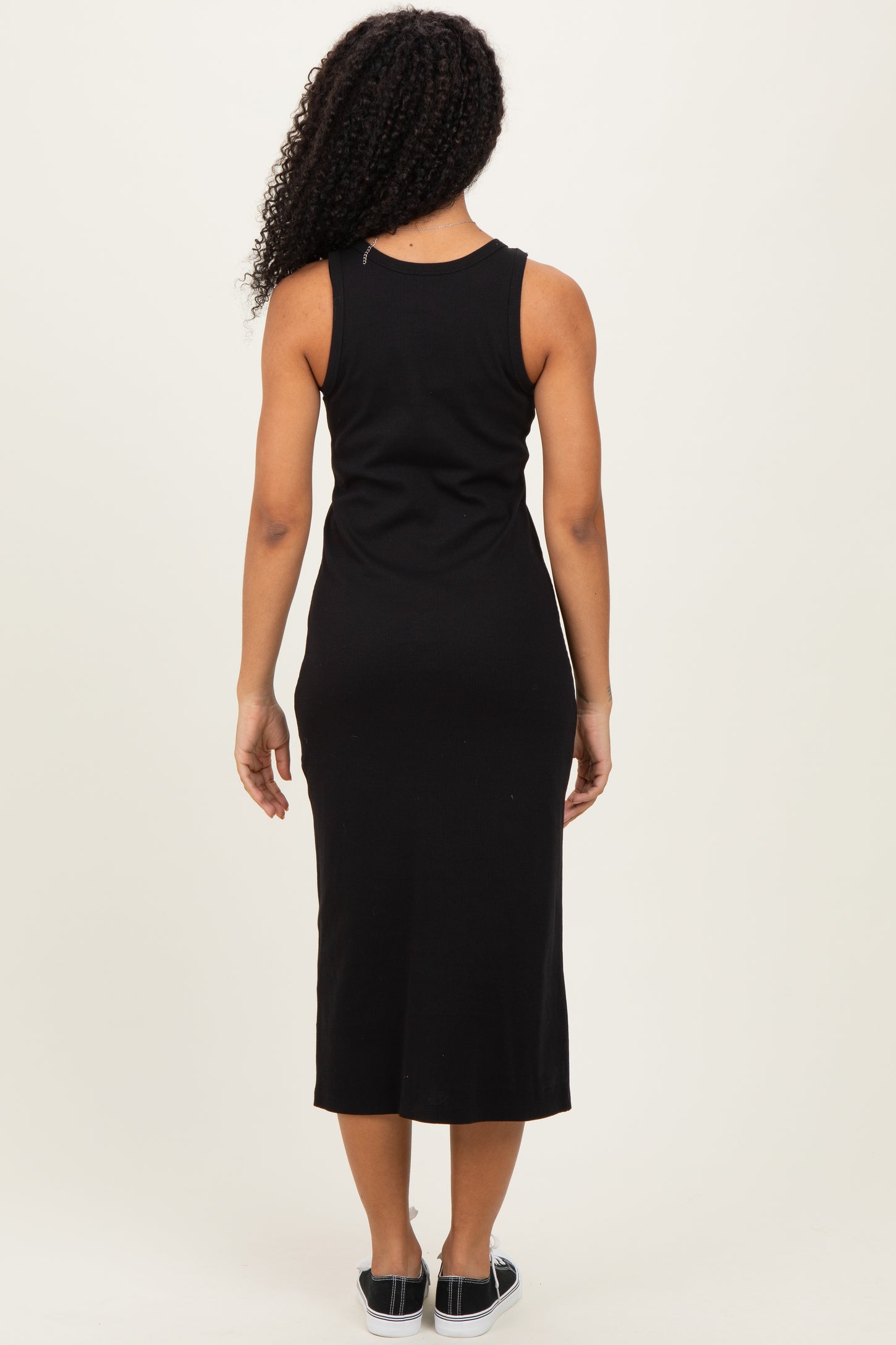 woman wearing a black sleeveless midi dress, shown from the back, with curly hair and casual footwear
