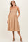 woman wearing a beige maternity dress with a smocked bodice and a full skirt, standing with hands in pockets
