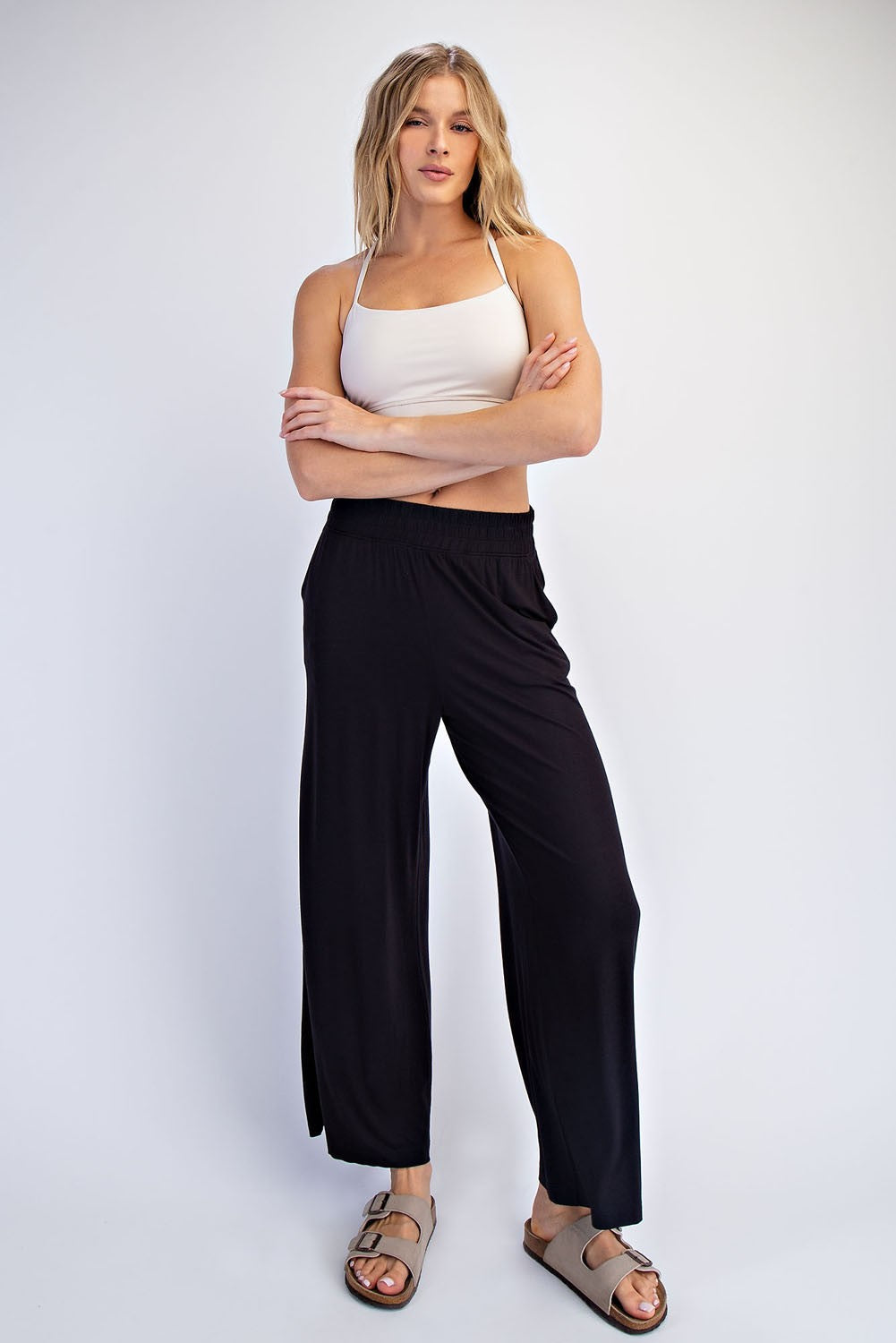 woman wearing a white tank top and black wide-leg pants, standing with arms crossed and wearing sandals