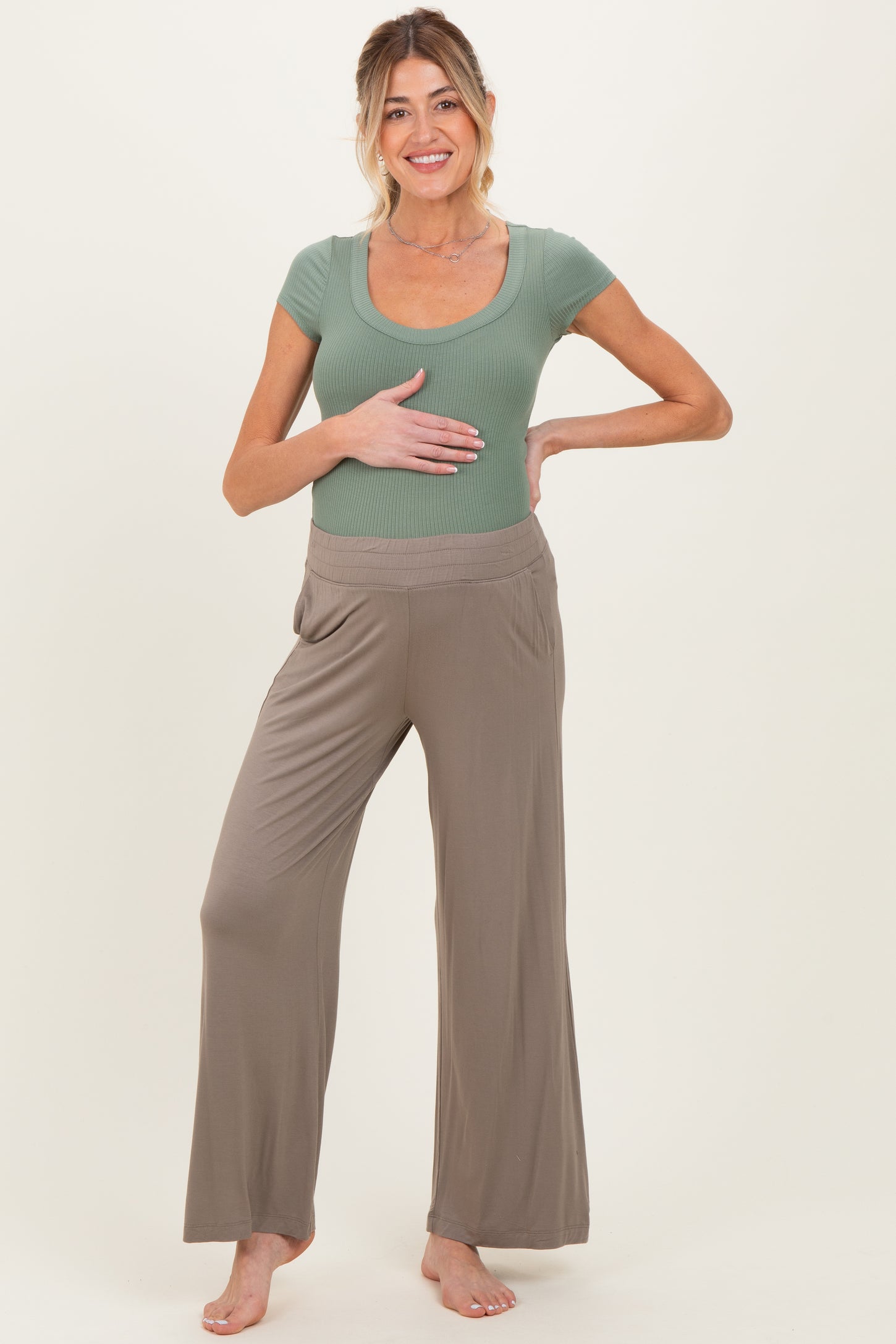 woman wearing a light green maternity top and beige maternity wide-leg pants while holding her baby bump
