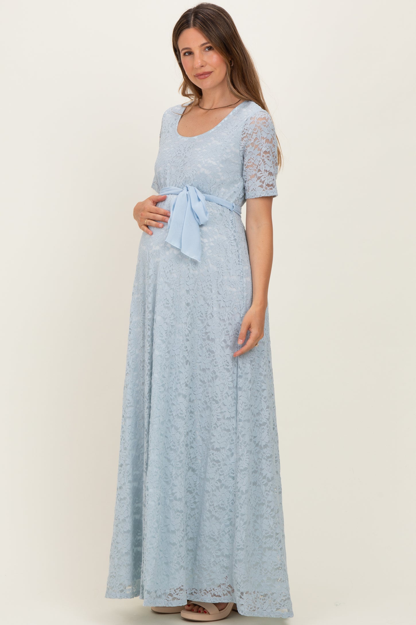 woman wearing a light blue lace maternity gown with a bow at the waist and holding her baby bump