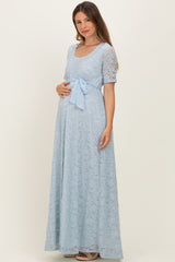 woman wearing a light blue lace maternity gown with a bow at the waist and holding her baby bump