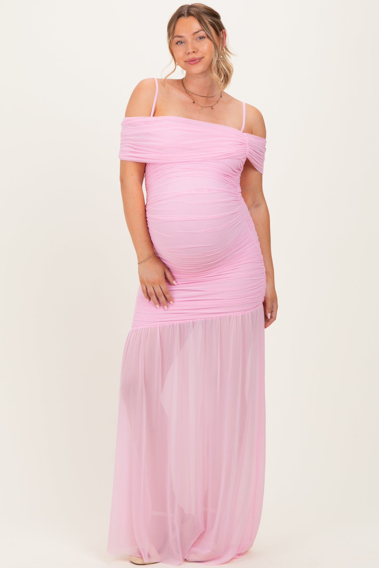 woman wearing a light pink off-the-shoulder maternity gown with ruched detailing and sheer skirt, showcasing her baby bump